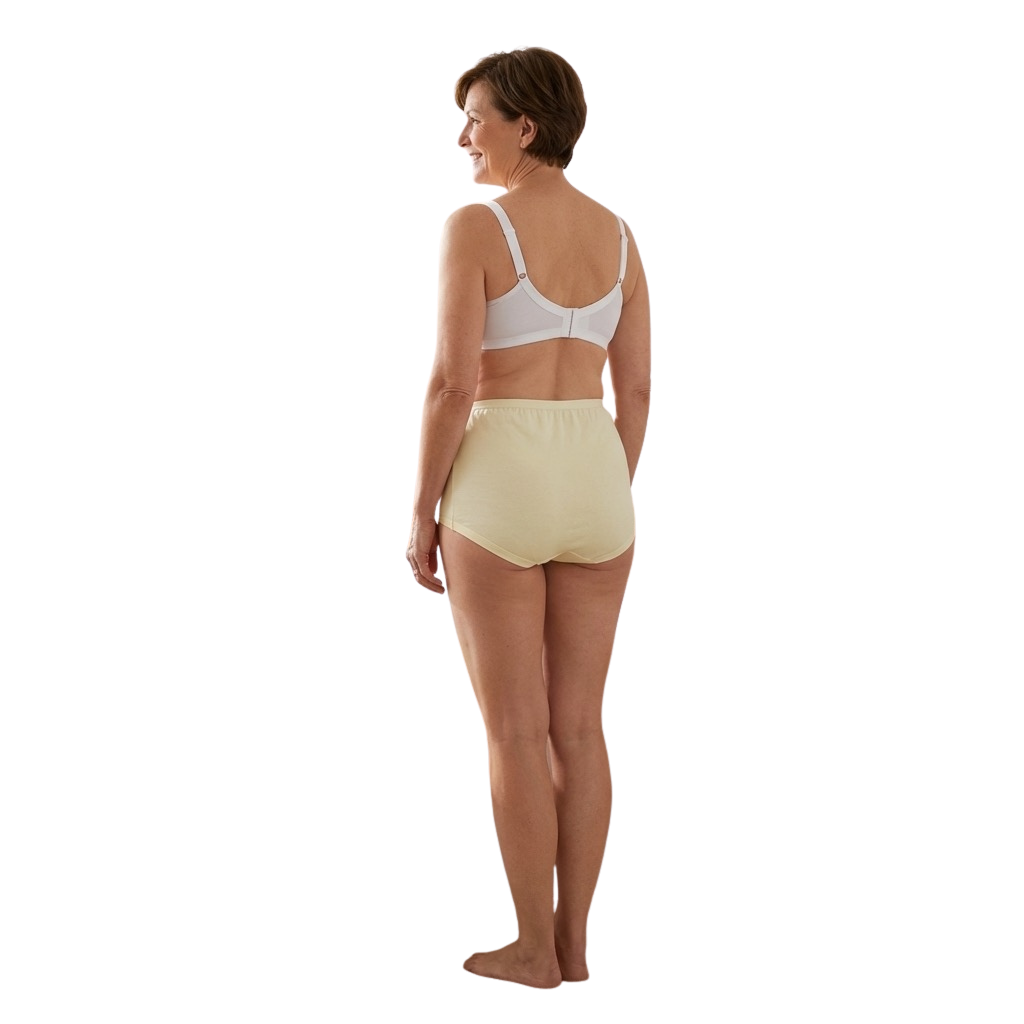 A woman with short brown hair stands barefoot, smiling and facing away in a white bra and Teri Lingerie's The Cotton Cuff Leg Betsy (M101), modest fit high-waist cotton panties in beige. The background is plain white.