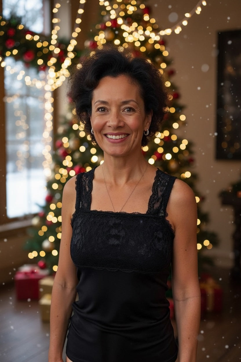 A woman with short curly hair, wearing a black lace top, smiles in front of a decorated Christmas tree with twinkling lights and ornaments. Snow appears to be falling inside the cozy room.